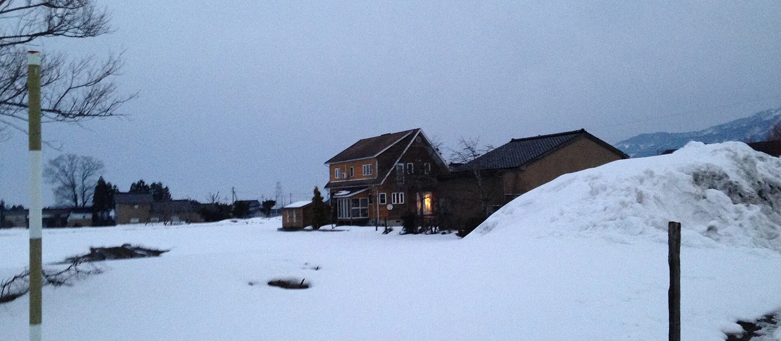 雪に覆われた田の向こうに、夕暮れの家並みと除雪した雪の山が見える南砺市城端の冬