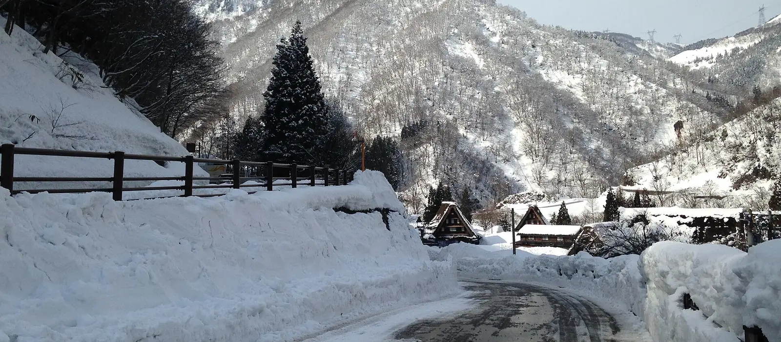 雪壁の間を抜けて菅沼集落へ下る道。奥に合掌造りの屋根（南砺市五箇山）